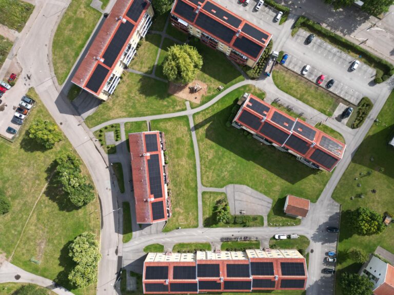 Flygfoto över flerfamiljshus med röda tak, gröna gräsmattor, parkeringsplatser och promenadstråk.