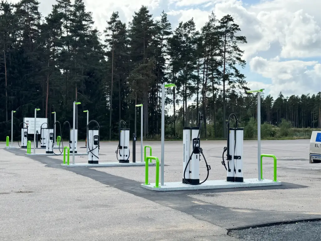 En rad laddningsstationer för elfordon står på en tom parkeringsplats nära ett skogsområde.