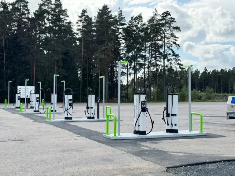 En rad laddningsstationer för elfordon står på en tom parkeringsplats nära ett skogsområde.
