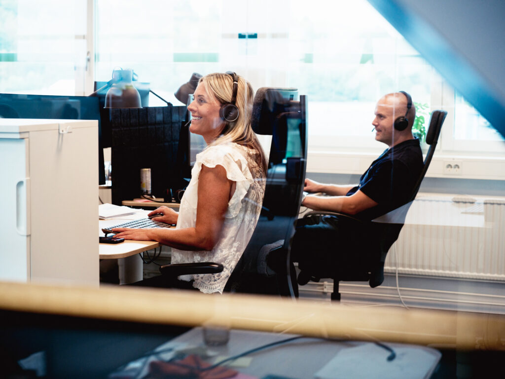 Två personer med headset sitter vid skrivbord med datorer i ett ljust, modernt kontor. Kvinnan i förgrunden ler och ger stöd, medan mannen i bakgrunden är fokuserad på sin skärm.