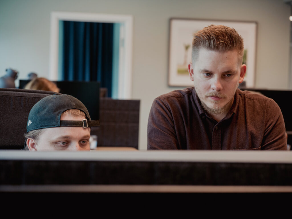 Två män arbetar tillsammans vid en dator på ett kontor och visar stöd när de fokuserar på bildskärmen. Den ene bär en bakvänd keps och sitter ner, medan den andre står bredvid honom. Kontorsinredning och andra människor syns i bakgrunden.