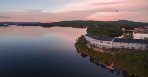 En stor byggnad ligger vid kanten av en lugn sjö i solnedgången, omgiven av träd och avlägsna kullar.