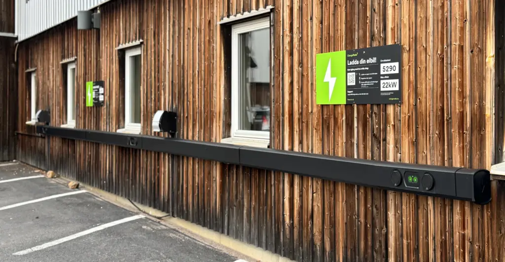Träpanelad byggnad med laddningsstationer för elfordon och en grön laddningsskylt på väggen.