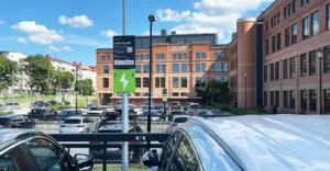 Parkeringsplats med många bilar, laddningsstationer för elfordon och en stor tegelbyggnad i bakgrunden.